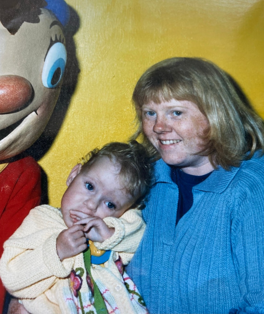 Young Danielle is pictured with her mum, posing next to Noddy. There is a yellow wall in the background. Danielle’s head is resting on her mum’s chest and she’s wearing a yellow cardigan with a green/pink pattern. She’s looking at the camera with a finger in her mouth. Her mum is wearing a blue top with a blue jacket, looking at the camera and smiling.
